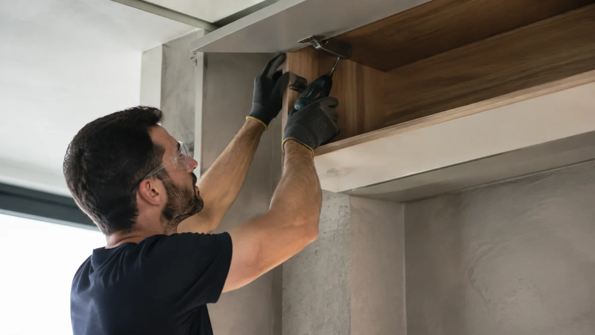 Profissional instalando ferragem em armário aéreo com uma parafusadeira ultracompacta e leve, braço elevado com postura natural e ferramenta pequena claramente visível